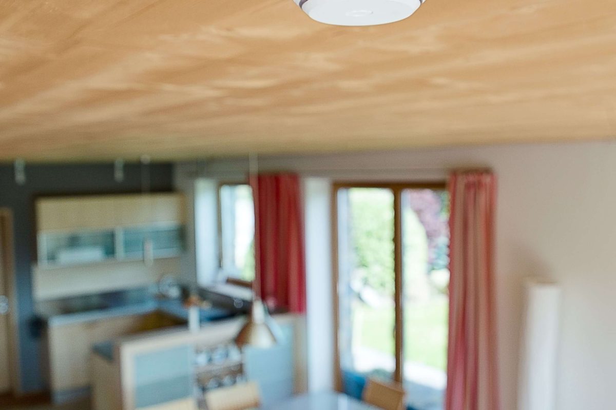 Rauchmelder an der Decke in einem modernen Wohnraum, im Hintergrund sind Küche und Essbereich zu sehen.