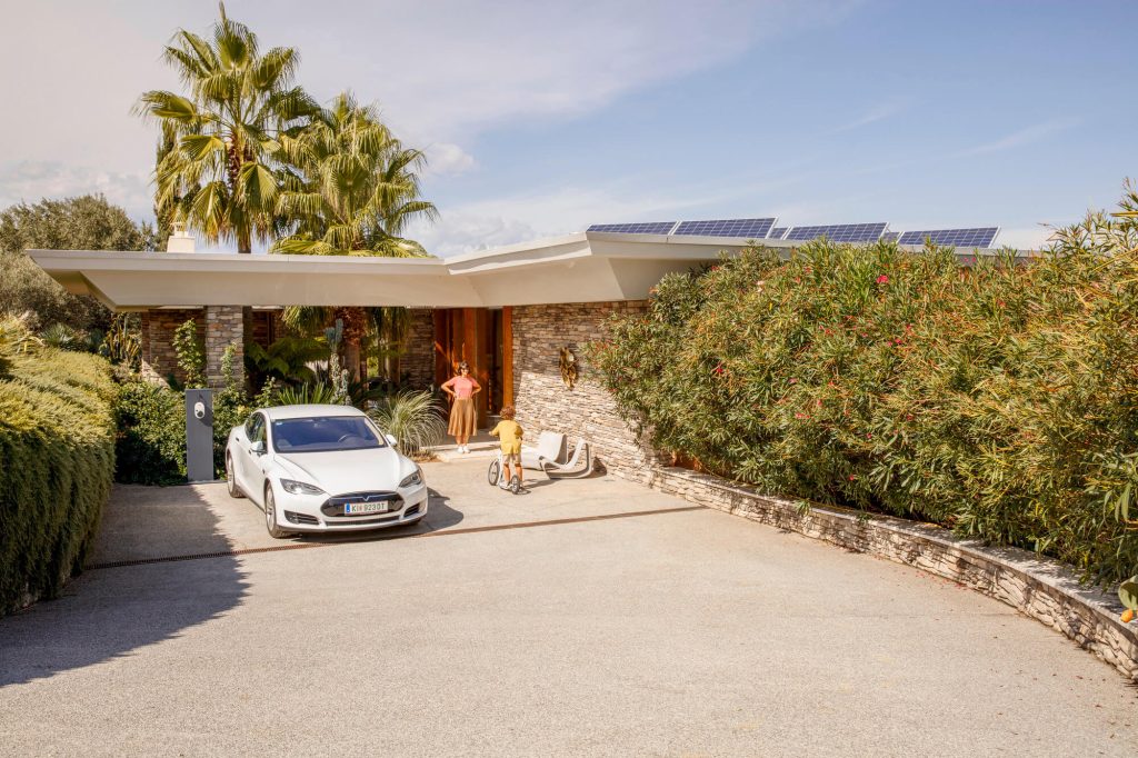Einfamilienhaus mit Solarpaneelen auf dem Dach, weißem Tesla vor der Garage und Frau mit Kind im Vorgarten.