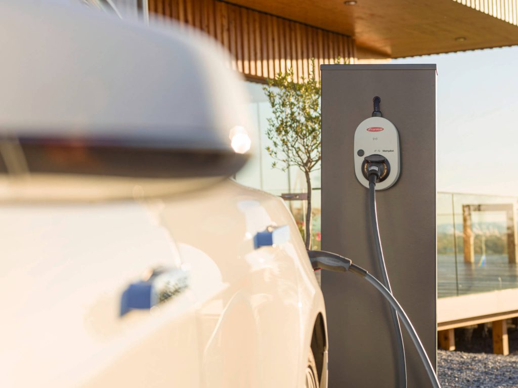 Weiße Wallbox mit angeschlossenem Ladekabel lädt ein weißes E-Auto vor einem modernen Haus.