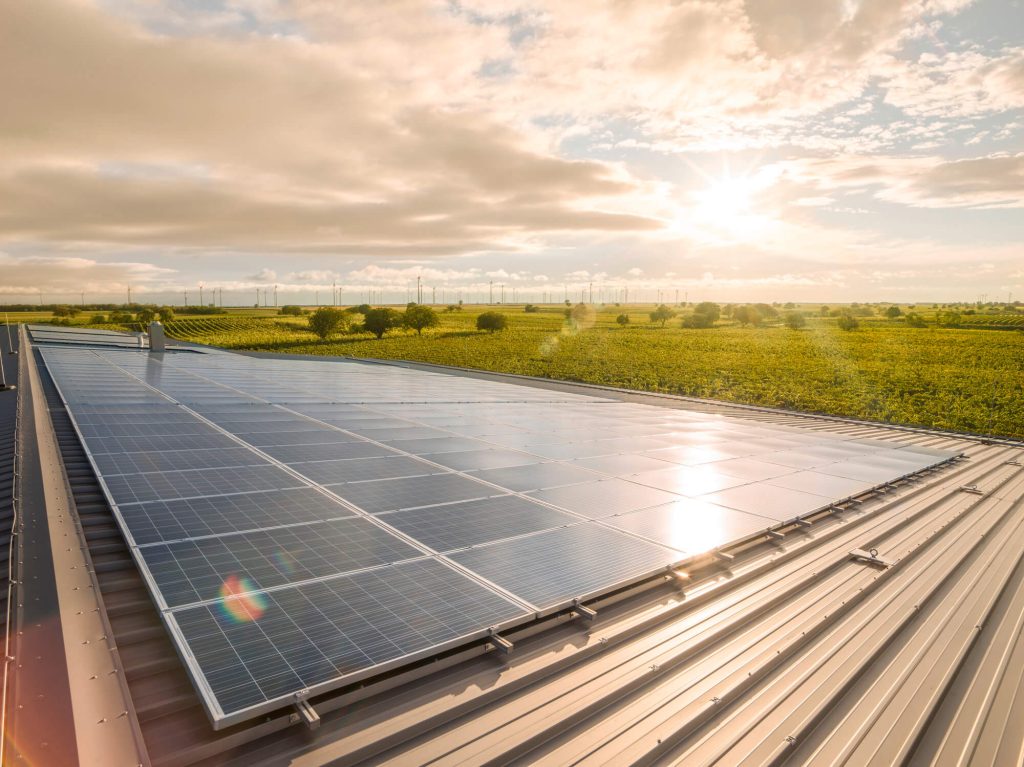 Große Solaranlage auf einem Hallendach mit Blick auf ein grünes Feld und Windräder in der Ferne bei untergehender Sonne.