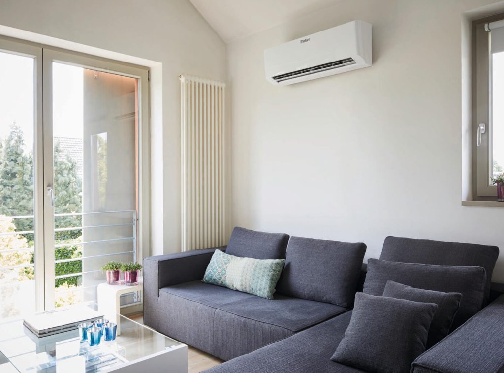Modernes Wohnzimmer mit Vaillant Klimaanlage an der Wand und vertikalem Heizkörper neben der Balkontür, eingerichtet mit grauem Sofa und Glas-Couchtisch.