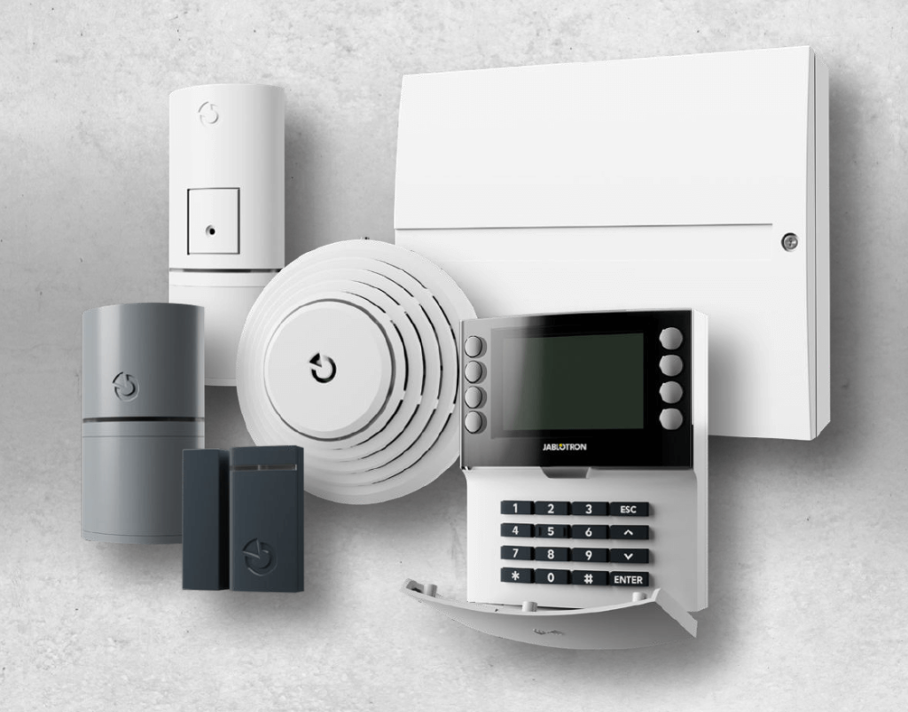 Komplettes Jablotron Alarmsystem bestehend aus Bedieneinheit, Bewegungsmeldern, Rauchmelder, Tür-/Fenstersensoren und Zentraleinheit vor grauem Hintergrund.