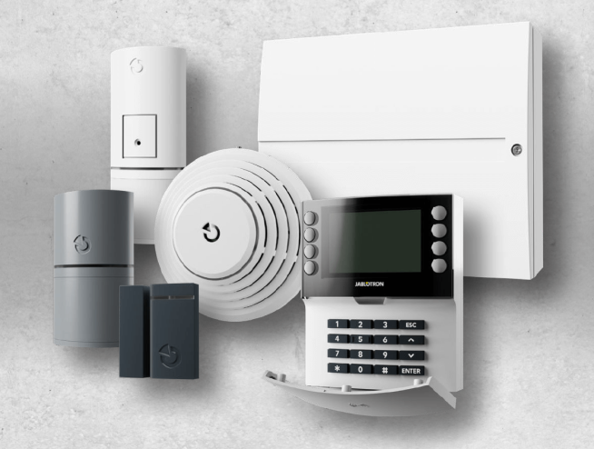 Komplettes Jablotron Alarmsystem bestehend aus Bedieneinheit, Bewegungsmeldern, Rauchmelder, Tür-/Fenstersensoren und Zentraleinheit vor grauem Hintergrund.