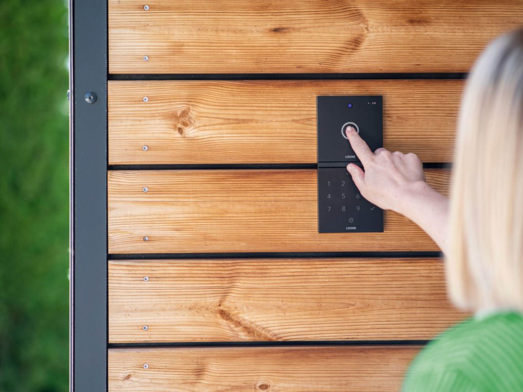 Hand einer Frau betätigt eine schwarze Loxone-Türstation mit Touchpanel an einer hölzernen Außenwand