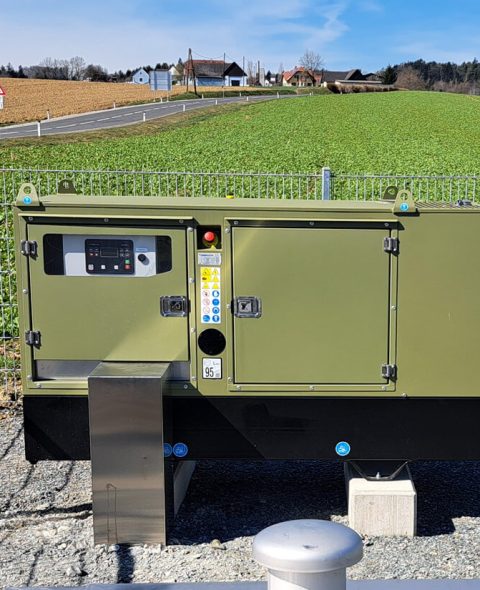 Olivegrünes Aggregat in einem umzäunten Bereich auf freiem Feld, dient als autarke Energiequelle